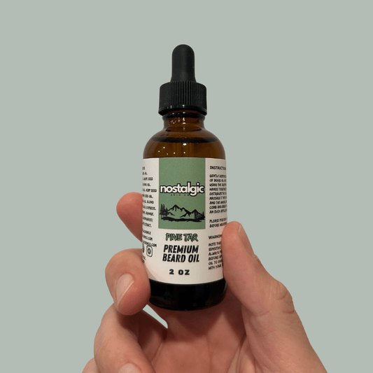 Hand holding a bottle of Nostalgic Skin Co Pine Tar Premium Beard Oil, 2 oz size, with green label and dropper cap on a muted green background.