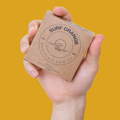 Hand holding Surf Orange men's bar soap packaging, displaying earthy design on a vibrant yellow background.