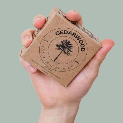 Hand holding Cedarwood bar soap packaging from Nostalgic Skin Co, featuring a tree logo on natural cardboard.